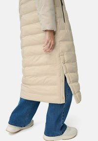Person iført en beige lang pufferjakke med knaplukning i siden, blå jeans og hvide sneakers, vist fra midt lår til fødderne mod en hvid baggrund.