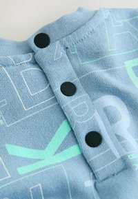 Svetlomodrá látka s geometrickými vzormi, so zaobleným výstrihom a tromi čiernymi gombíkmi vzadu na zapínanie.