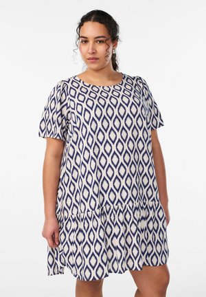 Kvinde med krøllet hår iført en kortærmet hvid kjole med blå geometriske diamantmønstre mod en ensfarvet baggrund.