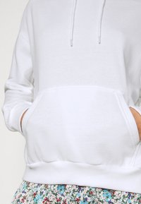 Personne portant un sweat à capuche blanc avec poches frontales et jupe à motif floral, les mains partiellement dans les poches du sweat.