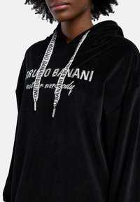 Schwarze Velours-Hoodie mit silberner bestickter Schrift und passenden Kordeln mit Branding. Weicher Stoff und lockere Passform.