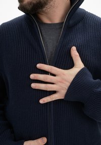 Marineblauwe gebreide ritsv sweater met een gestructureerde afwerking, voorzien van een hoge kraag en brede manchetten, gedragen over een donkergrijze shirt.