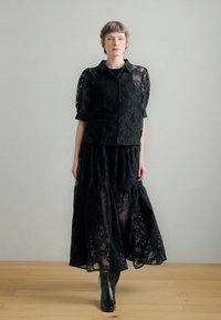 Μαύρο τοπ από δαντέλα με κοντά φουσκωτά μανίκια, συνδυασμένο με μακριά μαύρη φούστα από δαντέλα, και τα δύο με περίτεχνα σχέδια, φορεμένα με μαύρα μποτάκια αστραγάλου.