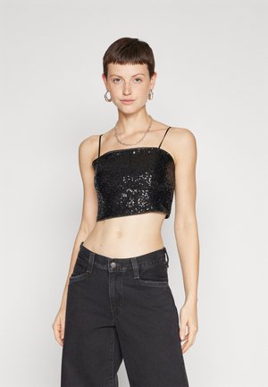 Νεαρή γυναίκα που φοράει μαύρο κεντημένο τοπ με λεπτές τιράντες, μαύρα τζιν ψηλής μέσης, ασημένιο κολιέ και γεωμετρικά σκουλαρίκια.