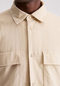 Beżowa koszula zapinana na guziki z teksturowanej tkaniny, z kołnierzykiem i dwoma kieszeniami na piersi z klapkami. Duże plastikowe guziki.