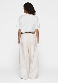 Mujer con cabello rizado que lleva una camiseta blanca metida en unos pantalones anchos color crema con grandes estampados de flores blancas y un cinturón oscuro, de espaldas.