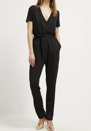 Femme portant une combinaison noire à manches courtes avec un col en V croisé et une ceinture à la taille, associée à des talons hauts métalliques à bouts pointus.