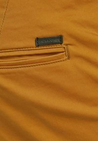 Gros plan d'un tissu couleur moutarde avec une petite étiquette noire cousue au-dessus d'une poche fente horizontale.