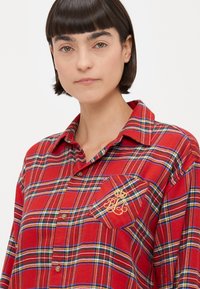 Camicia a quadri rossi in tessuto morbido. Presenta un design con bottoni, una tasca con dettagli ricamati e strisce blu e verdi a contrasto.