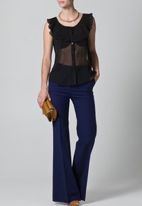 Blusa nera a plissettature con collare volante, chiusura con bottoni, abbinata a pantaloni navy a gamba larga e una pochette marrone. Texture lisce e design aderente.