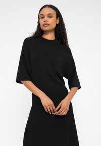 Fekete kötött felső rövid ujjakkal és kerek nyakkivágással. Az anyag puha tapintású, úgy van kialakítva, hogy a derék része tenyérszorító, és folyékony sziluettet ad.