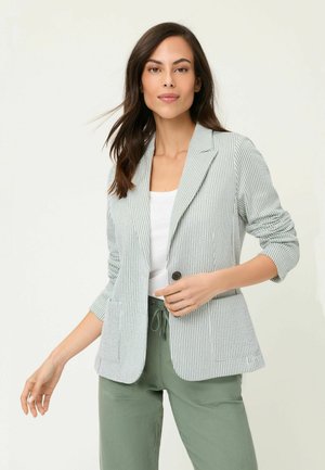 Donna che indossa una giacca a righe grigio chiaro sopra una maglietta bianca e pantaloni verde oliva, posando con una mano vicino al bottone della giacca.