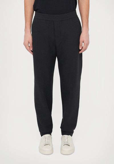 Black sweatpants with a textured fabric, elastic waistband, and tapered leg design, paired with light-colored sneakers.
