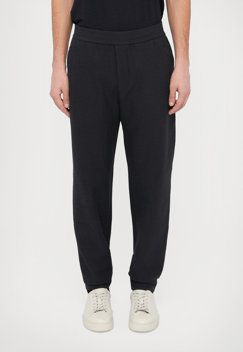 Czarne spodnie dresowe z fakturowanej tkaniny, elastycznym pasem i zwężanymi nogawkami, zestawione z jasnymi sneakersami.