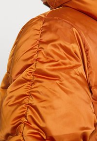 Puffjacka i livlig orange satin, med rynkad textur längs sömmen för extra detalj och en mjuk finish.
