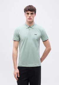 Νέος άνδρας με κοντά σγουρά μαλλιά, φορώντας ανοιχτό πράσινο πόλο με το λογότυπο «BOSS» και μαύρο παντελόνι, στέκεται μπροστά σε απλό λευκό φόντο.