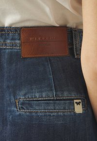Jeans de mezclilla con un parche de cuero marrón en la cinturilla etiquetado "WEEKEND MaxMara", que cuenta con un bolsillo trasero y una pequeña etiqueta de la marca con un logo de mariposa.