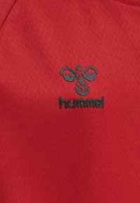 Rød stofskjorte med en glat tekstur, der har et sort Hummel-logo formet som en bi på venstre side.