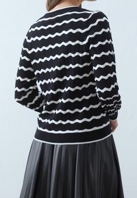 Randig svart och vit tröja med ett vågigt mönster, rund hals, långa ärmar och en åtsittande fåll, tillsammans med en grå plisserad kjol.