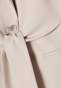 Gros plan sur un blazer beige avec une ceinture en tissu nouée, un bouton rond unique et une poche fendue sur un tissu à texture lisse.