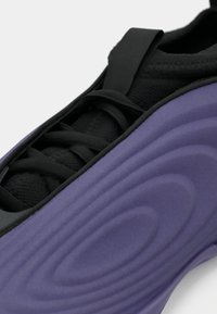 Chaussure de sport avec une tige en mesh noir et un revêtement en caoutchouc violet texturé. Comprend des lacets noirs et un design épuré avec des détails de contour.