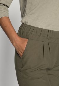Pantalons vert olive avec une taille élastique, dotés de poches latérales et de plis, fabriqués dans un matériau lisse et extensible.