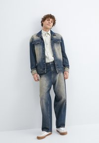 Denimjakke med falmede blå og lysebrune nuancer, parret med matchende falmede jeans. Hvid button-up skjorte under, hvide sneakers med brune såler.