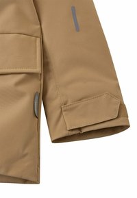 Beige wasserdichte Jacke mit Ärmeln, die über einen Klettverschluss verfügen, seitlichen Taschen und grauen reflektierenden Akzenten. Glatte Textur mit gesteppten Details.