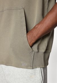 Sweatshirt verde oliva claro com um bolso frontal e punhos canelados, combinado com calças de moletom cinzentas com um pequeno detalhe de logo perto da bainha.