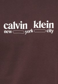 Σκούρο καφέ βαμβακερό μπλουζάκι με άσπρο κείμενο που γράφει «calvin klein new york city». Η γραμματοσειρά συνδυάζει κεφαλαία και μικρά γράμματα.
