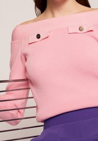 Kvinna klädd i en ribbade rosa off-shoulder tröja med metallknappar, i kombination med högmidjade lila byxor, nära en metallstolsgavel.