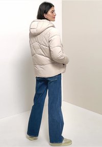 Beige, kviltet puffer-jakke med hætte, der har en tekstureret overflade og vandrette sømme, parret med blå, vidde jeans og lyse sneakers.