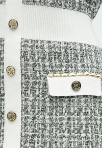 Πλεκτό ύφασμα σε γκρι και λευκό, με μεγάλες χρυσές κουμπιά και τσέπη με χρυσή αλυσίδα. Υφή με κάθετα σχέδια.
