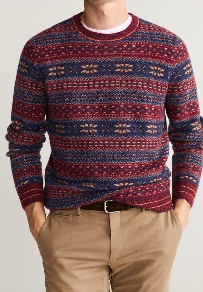 Homme portant un pull en tricot rouge et bleu à motifs, un pantalon beige et une ceinture noire, les mains dans les poches, debout devant un fond uni.