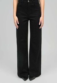 Femme portant un pantalon noir large taille haute à texture à petits pois, associé à des chaussures noires à bout pointu, se tenant devant un fond uni.