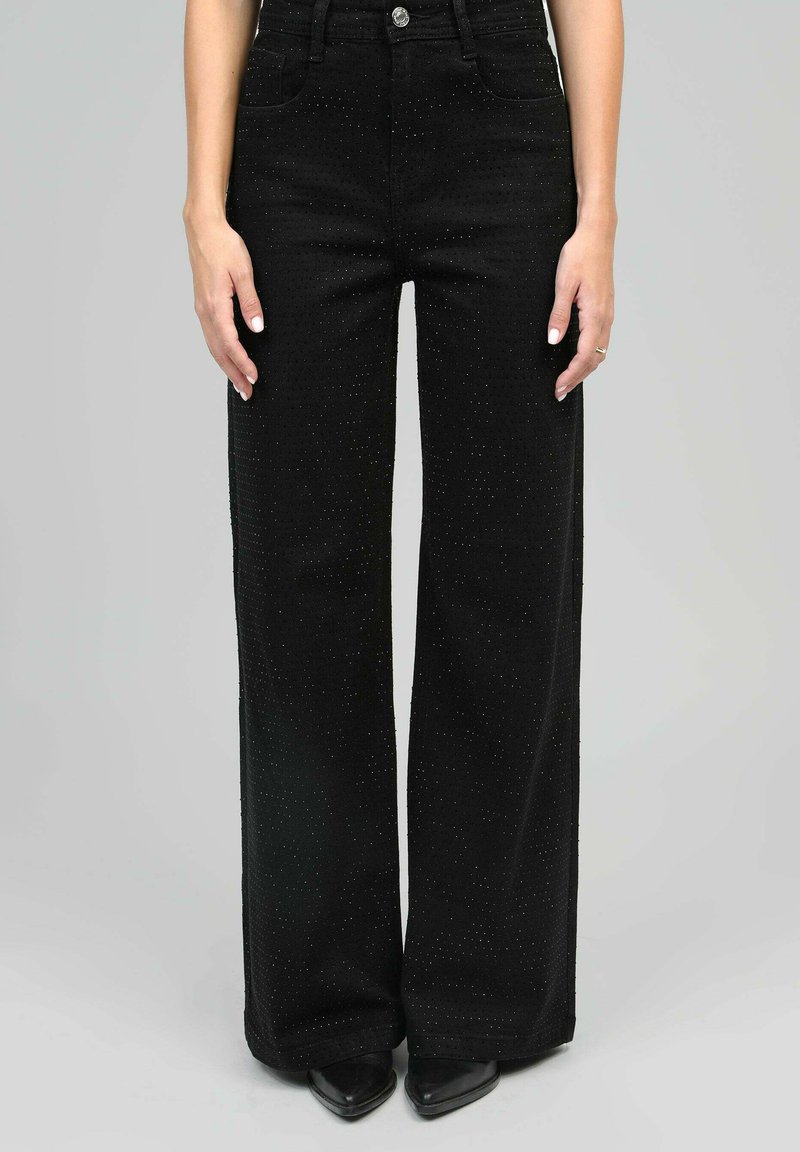 Femme portant un pantalon noir large taille haute à texture à petits pois, associé à des chaussures noires à bout pointu, se tenant devant un fond uni.