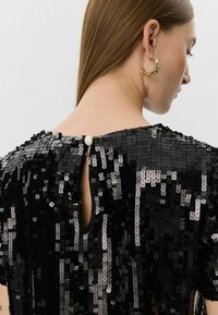 Sort glitrende kjole med rund halsudskæring, detalje med nøglehul i ryggen og lukning med perleknap. Glat tekstur med reflekterende detaljer.