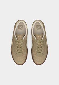 Ζευγάρι μπεζ παπουτσιών Veja με ανοιχτό κίτρινα στοιχεία, λεπτομερείς ραφές και καστανές λαστιχένιες σόλες, απόψη από ψηλά σε λευκό φόντο.