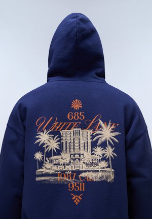 Bagudvendt billede af person iført marineblå hættetrøje med beige palmetræsprint, bygningsillustration og orange tekst "685 White Lane 1987 CAL 9511."