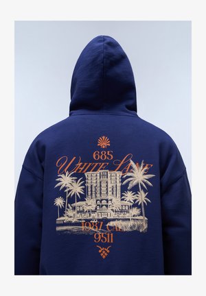 Henkilön takaosa, joka pukeutuneena laivastonsiniseen huppariin, jossa on beigenvärisiä palmupuita, rakennuksen kuva ja oranssi teksti "685 White Lane 1987 CAL 9511."