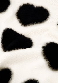 Gebreid sweater met een witte basis, versierd met grote zwarte fuzzy hartvormen die over het oppervlak verspreid zijn, wat een textuurpatroon creëert.