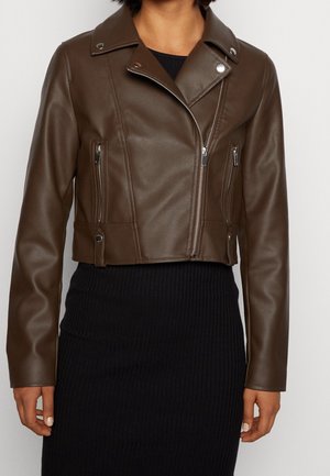 Femme portant une veste de motard en cuir marron courte avec des fermetures éclair argentées sur une robe noire côtelée, montrée des épaules aux hanches.