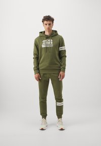 Grüne Kapuzenjacke und passende Jogginghosen aus weichem Stoff. Mit weißem Text und Streifen. Rundhalsausschnitt, verstellbare Kapuze und anliegende Bündchen.