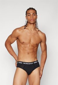 Schwarze Baumwoll-Slips mit einem elastischen Bund, der weiße "WORKSHOP 032c"-Markenaufdrucke zeigt. Enganliegendes Design und glatte Textur.