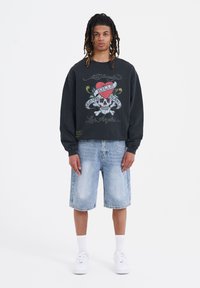 Svart sweatshirt med färggrafik av döskalle och hjärta, kombinerad med ljusblå denimshorts. Vita sneakers och ankelsockor fulländar outfiten.