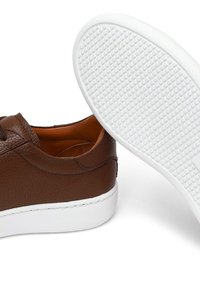 Bruine leren sneakers met een witte rubberen zool, gestructureerd oppervlak en gestikte details. Zijkant toont het interieur en een gedeelte van de zool.