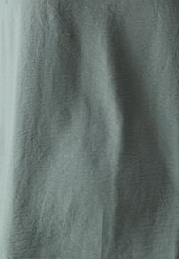 Gros plan sur un tissu gris clair avec de légères rides et une texture subtile, montrant des plis naturels et des coutures au bord inférieur.