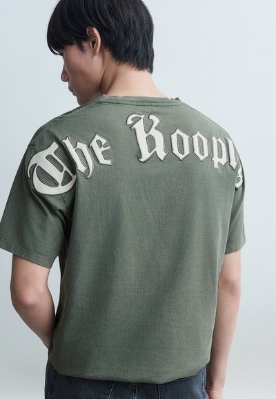 Jeune homme de dos portant un t-shirt vert olive avec un grand texte gothique blanc sur le haut du dos, sur un fond uni.