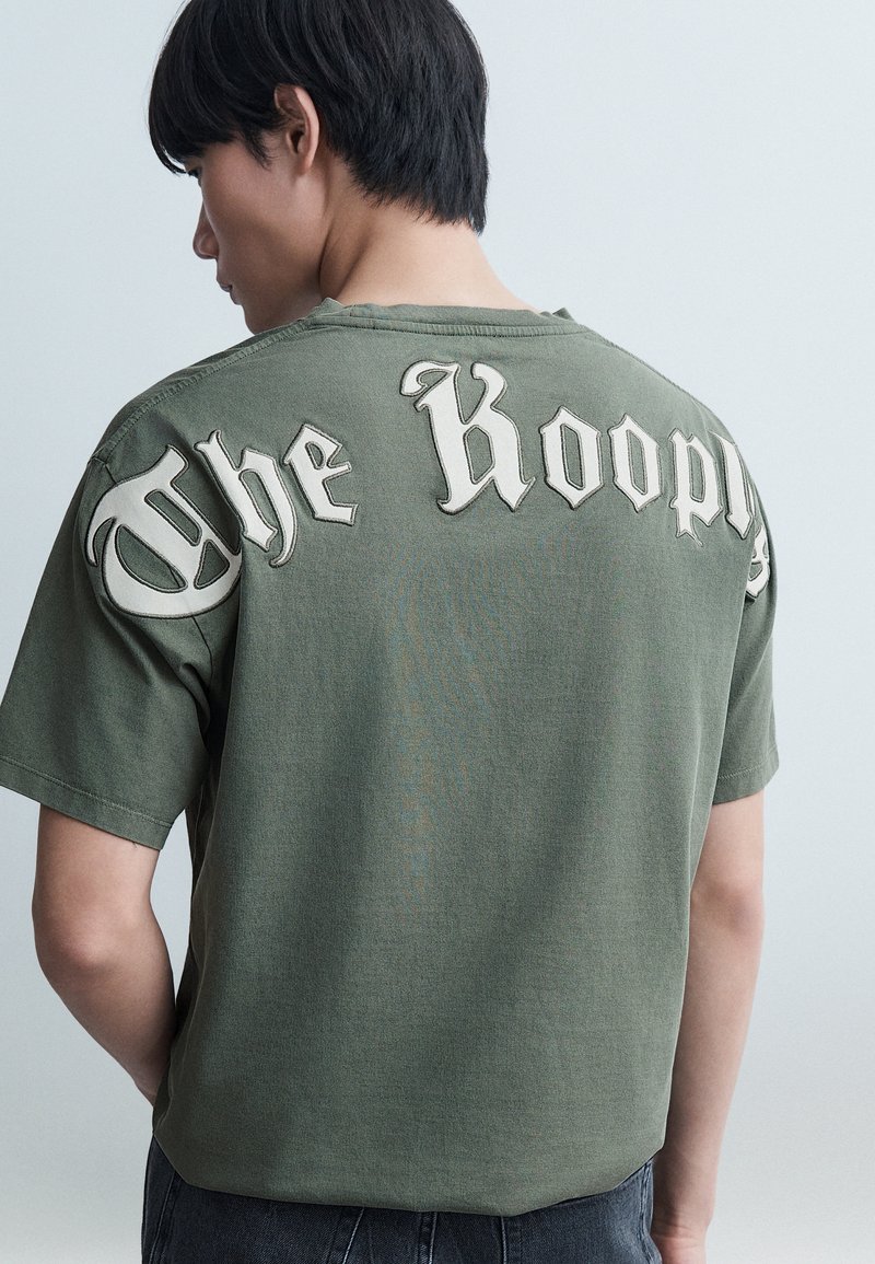 Νέος άνδρας που φαίνεται από πίσω φοράει χακί πράσινη μπλούζα με μεγάλο λευκό γοτθικό κείμενο στο πάνω μέρος της πλάτης, πάνω σε απλό φόντο.