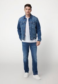 Denim jack in medium blauw met knoopsluiting, twee borstzakken en een puntige kraag. Draag het over een witte t-shirt en blauwe jeans. Witte sneakers.